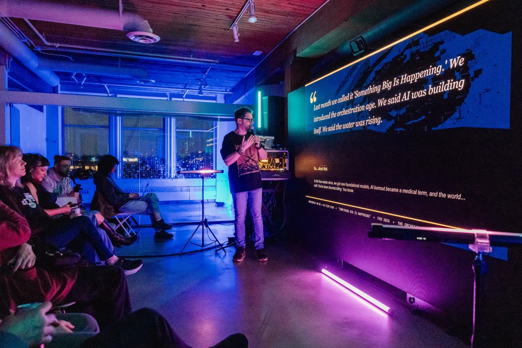 A speaker addresses a seated audience beside a large projection screen at Multimodal Media Lab.