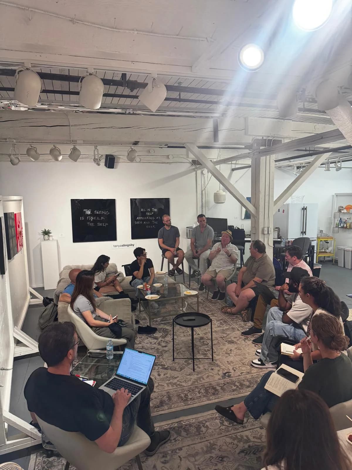 About twenty people seated in a circle of couches and chairs at Parker Street Studios for AI Ethical Futures Lab number 6, with a laptop open in the foreground and paintings on the gallery wall.