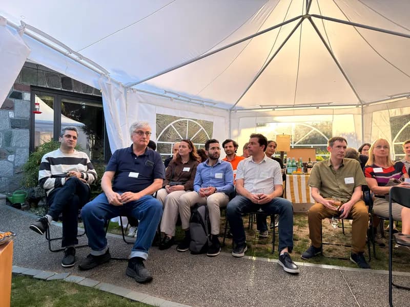 A row of attendees seated along the garden path under the tent, listening, glasses in hand.