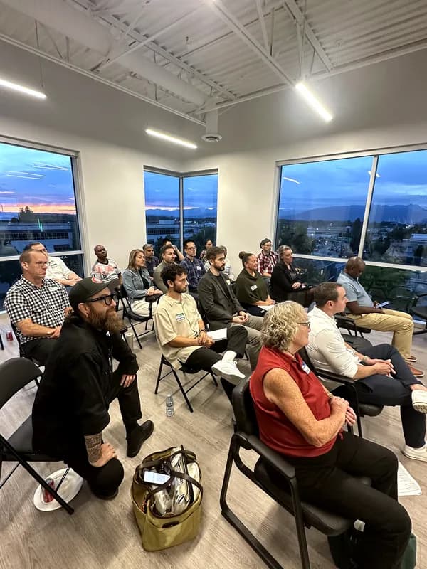 Attendees listen from folding chairs as the sunset fades to blue across three walls of windows.