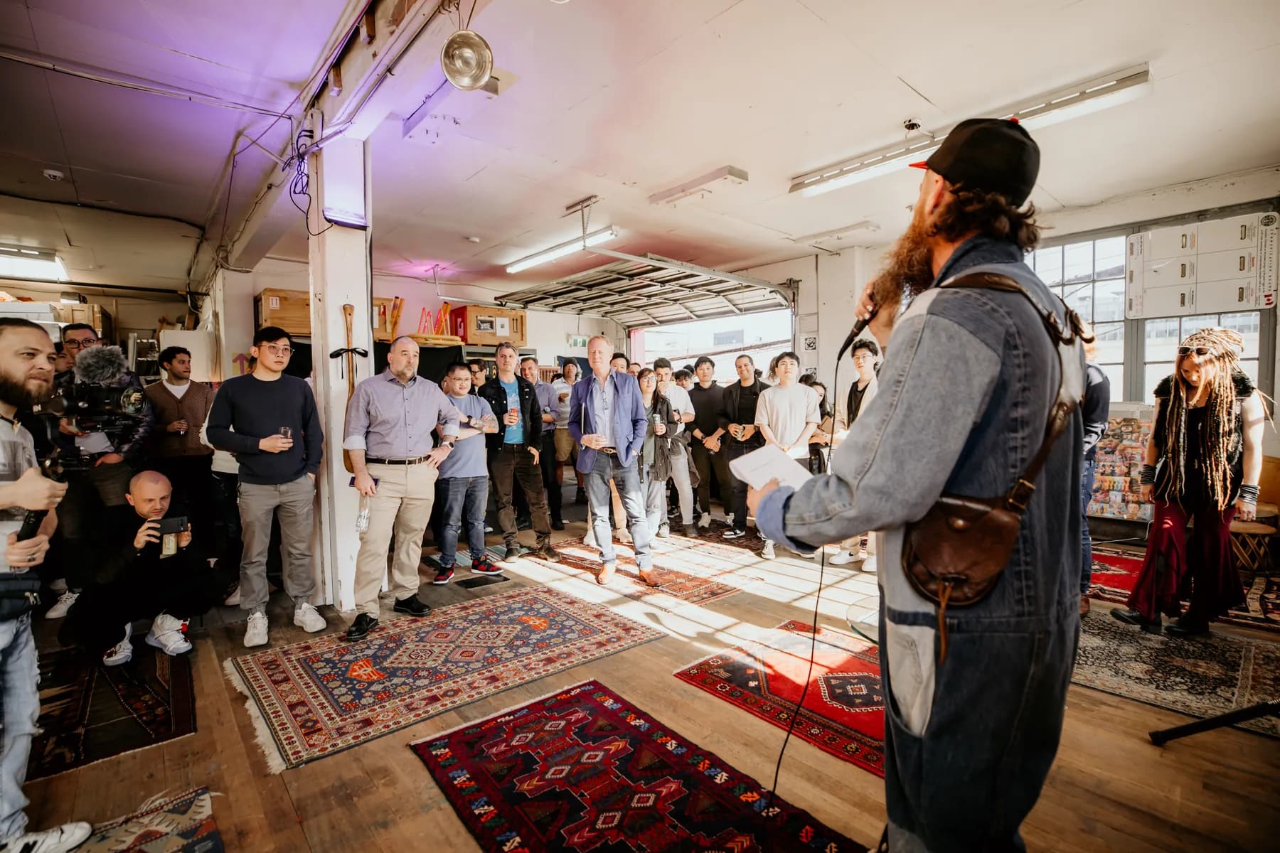 Kris Krüg facing a packed standing-room loft, notes in one hand and a microphone in the other, a camera operator filming from the left.
