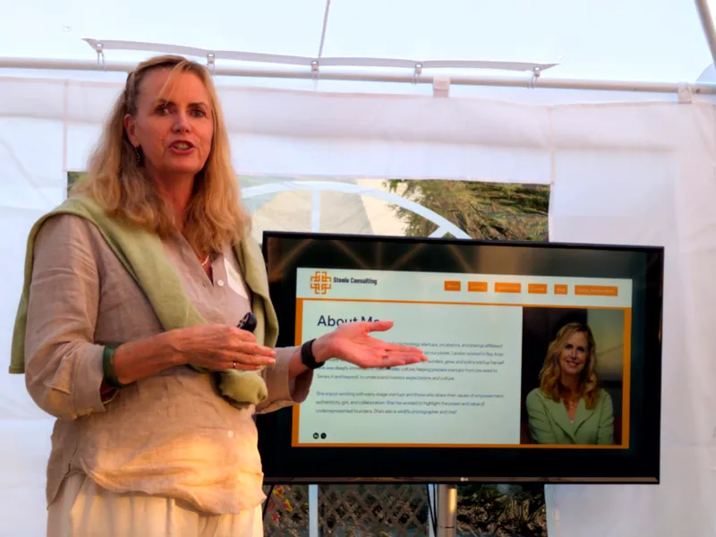 A speaker presenting beside a screen showing her consulting practice's About page.