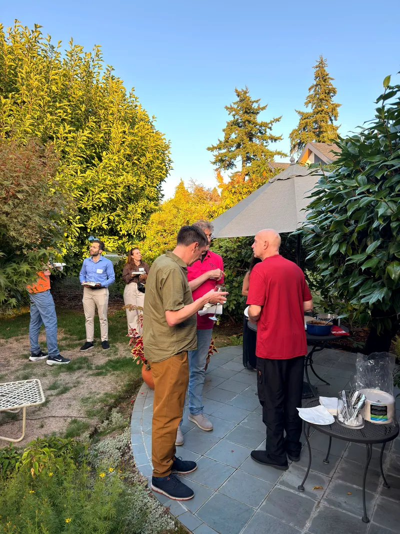 Guests standing on the back patio in low evening light, plates in hand, garden and firs behind.