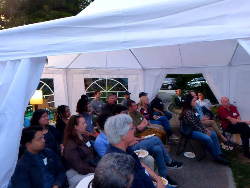 The group seated under the tent at dusk, a lamp lit at the left, plates balanced on knees.