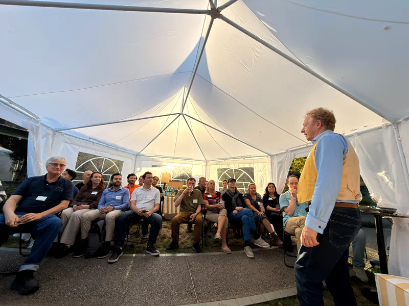 The full group seated under the marquee canopy as the host addresses them, low sun through the tent walls.