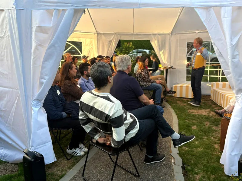 The seated group viewed from the tent entrance, curtains framing the shot, the host speaking at right.