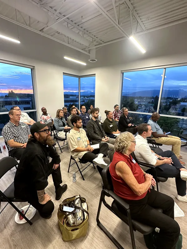 Attendees listen from folding chairs as the sunset fades to blue across three walls of windows.