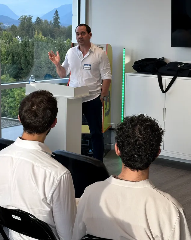 Darren stands at a podium welcoming the room to the first FV+AI meetup, with the Fraser Valley visible through the windows behind him.