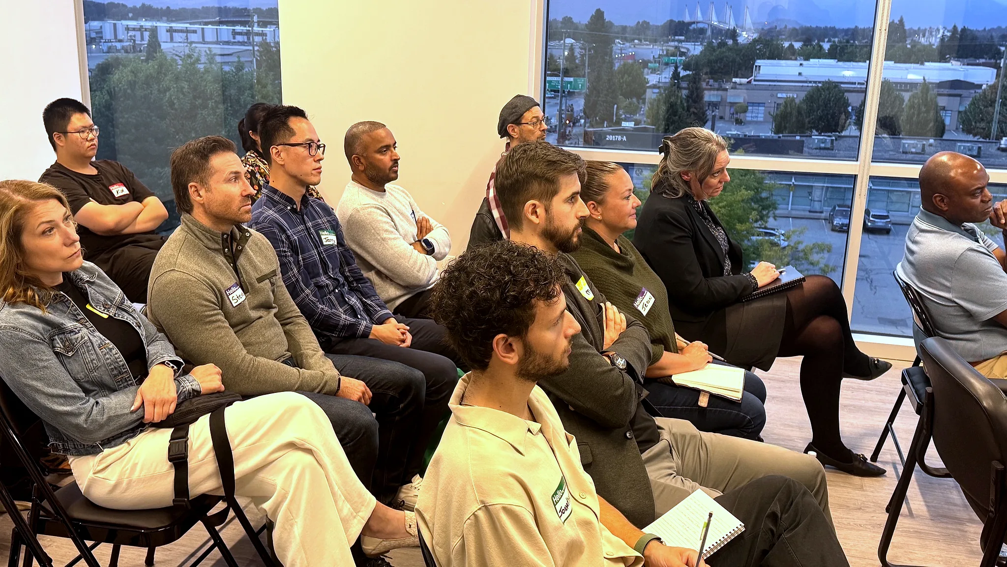 Attendees seated in rows of folding chairs listen to a talk, with the Fraser Valley and the Port Mann bridge visible through the windows at dusk.