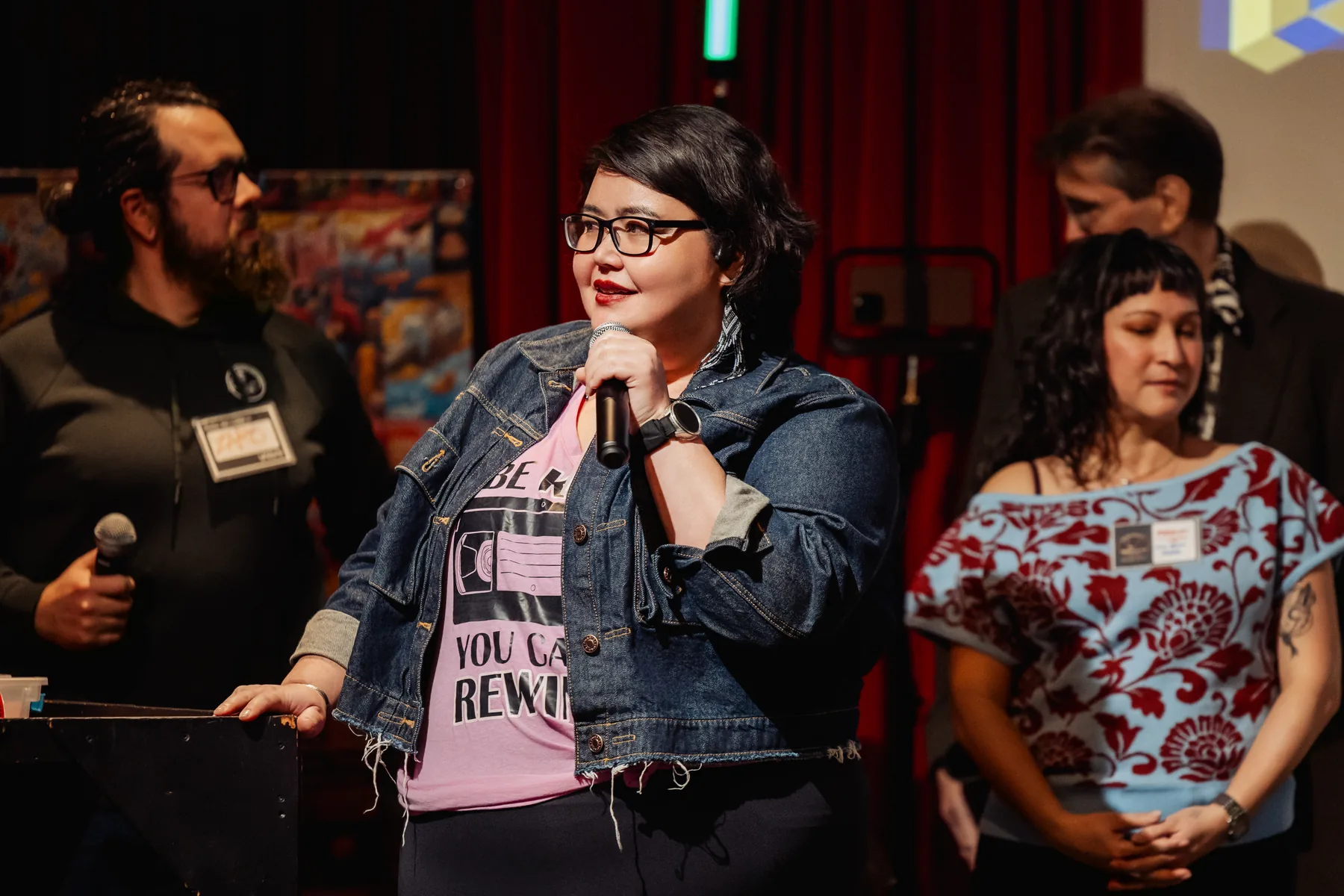A community member standing on stage with a microphone, mid-sentence, other members waiting their turn beside her.