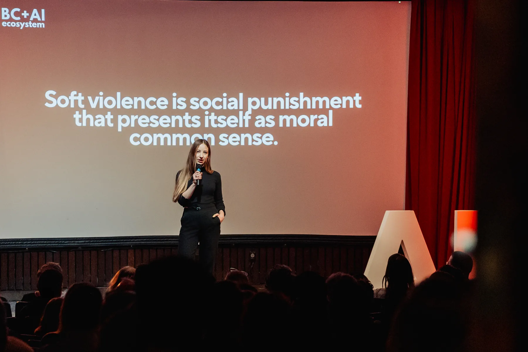 A speaker on stage in front of a large screen displaying a quote about soft violence and social punishment.