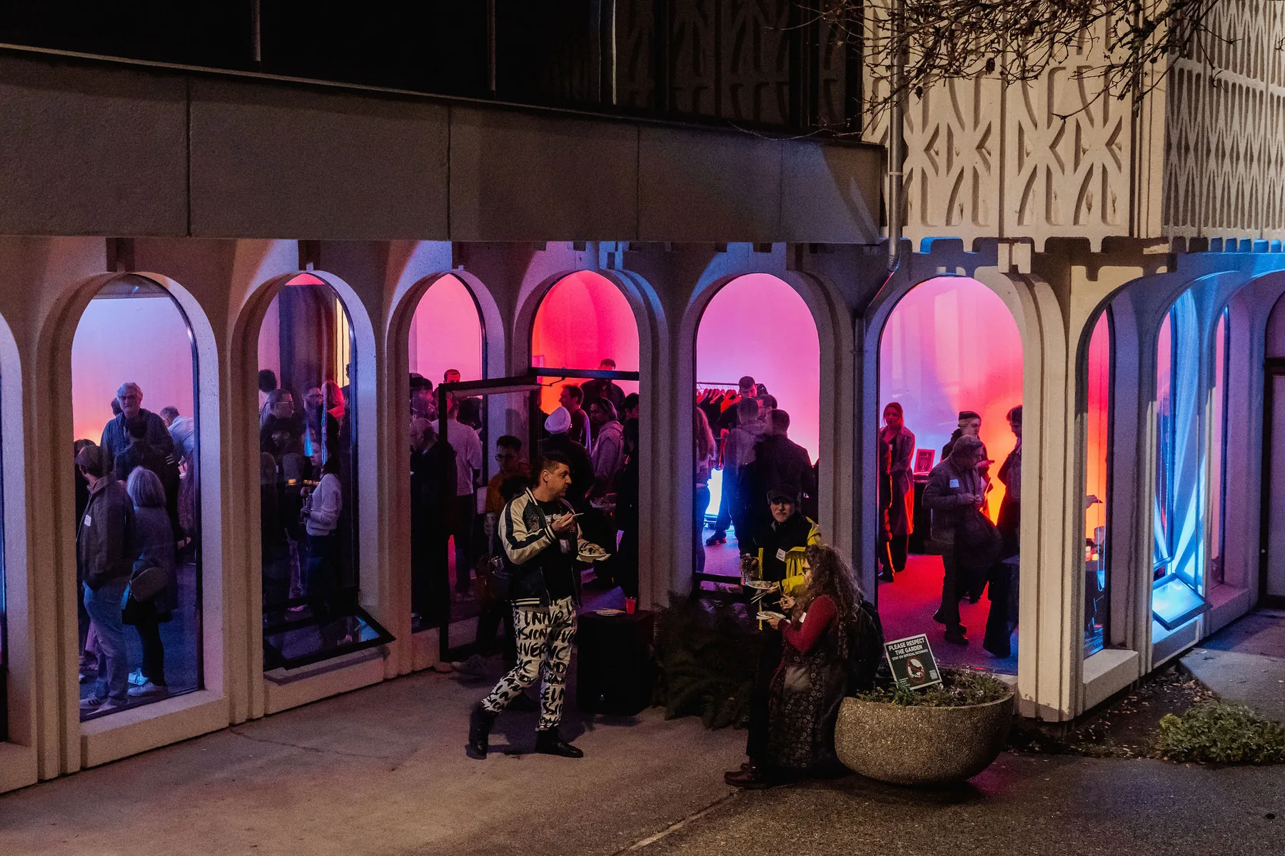 People talking and sharing food around arched windows at an evening community gathering, with pink and blue light inside.