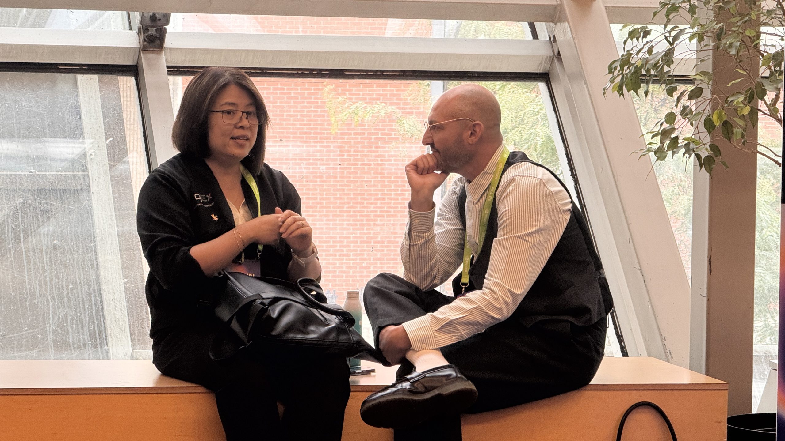 Two conference attendees seated beside a window, talking away from the ALL IN expo floor.