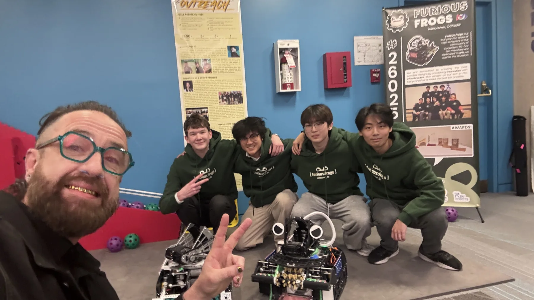 Kris Krug and Furious Frogs students smile with two competition robots and a team banner.
