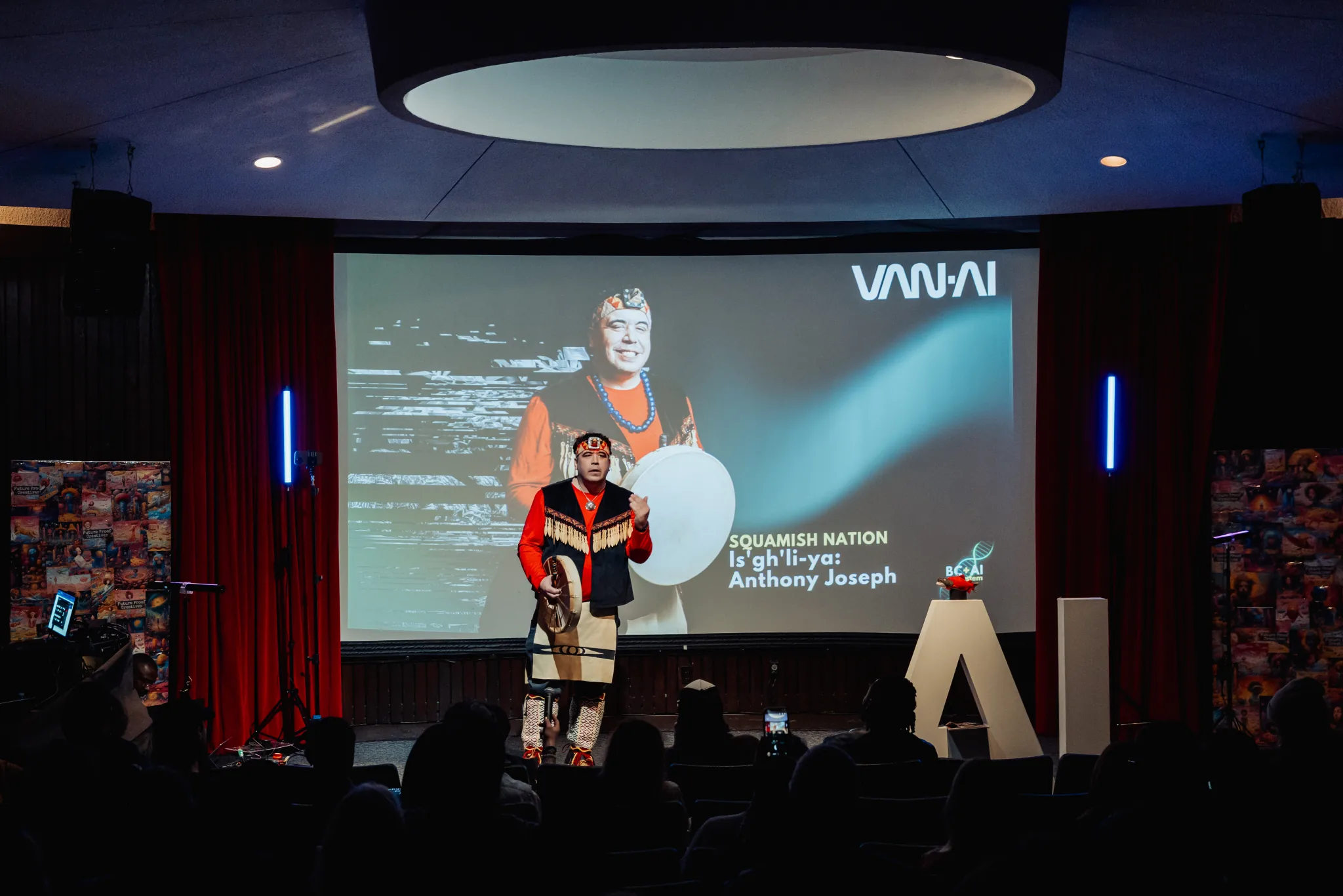 Anthony Joseph of Squamish Nation stands on the BC + AI stage with a drum.